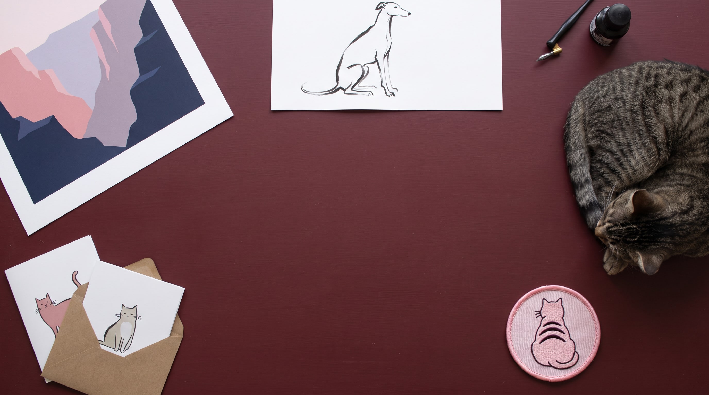 Art prints, folded greeting cards and an embroidered striped-cat patch on an oxblood table, a tabby cat asleep at the edge
